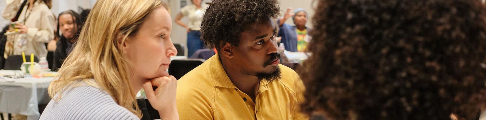 White womand and black man sit, listening to someone off camera speak