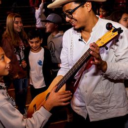 A mexican man shows a guitar to a young bow in front of a crowd.