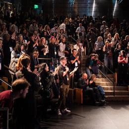An older woman speaks to a crowded auditorium